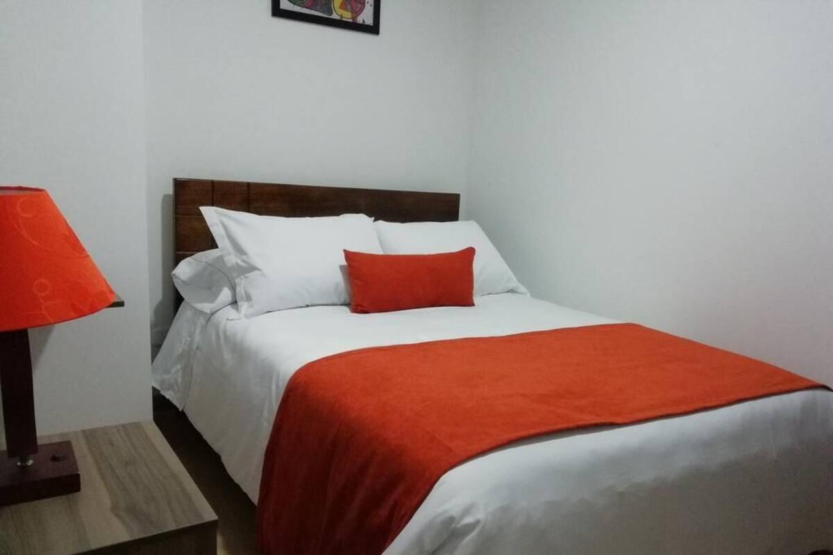 A neatly made bed with white and red bedding, a wooden headboard, and a small table with a red lamp beside it.