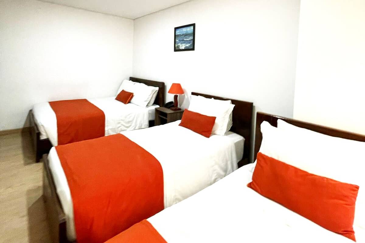 A hotel room with three beds, each with white and orange bedding. There is a small table with a lamp between the beds.