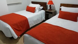 Dos llits individuals amb roba de llit blanca i vermella, una tauleta de nit amb una làmpada i una finestra amb persianes.