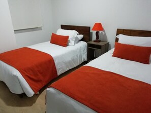 Dos llits individuals amb roba de llit blanca i vermella, una tauleta de nit amb una làmpada i una finestra amb persianes.
