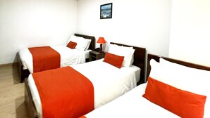 Habitació d'hotel amb tres llits, cadascun amb roba de llit blanca i taronja. Hi ha una taula petita amb una làmpada entre els llits.