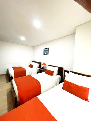 Quarto de hotel com três camas, cada uma com roupa de cama branca e laranja. Há uma pequena mesa com abajur entre as camas.