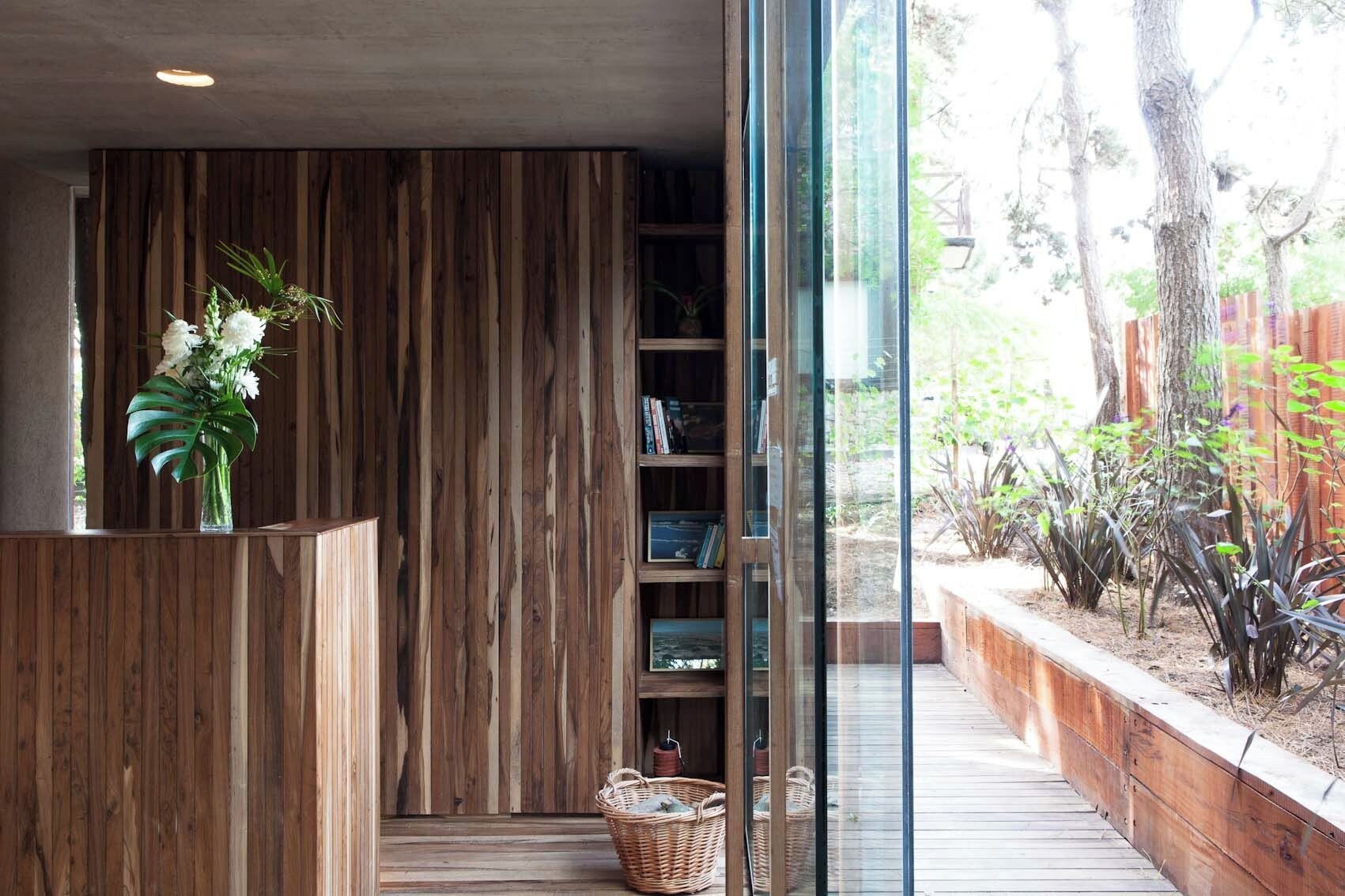 A wooden reception desk with a vase of flowers, a bookshelf with books, and a view of a garden through a glass door.