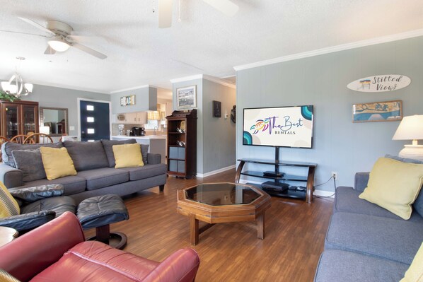 A living room with a sofa, ottoman, and glass coffee table. There is a television on a stand, a ceiling fan, and a kitchen in the background.