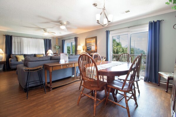 A living room with a sofa, dining table, chairs, and a balcony with curtains. - Across from Beach w/Lighthouse View!~Stilted Views (Folly Beach)