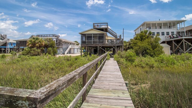 A wooden boardwalk leading to a house with a balcony, surrounded by vegetation.