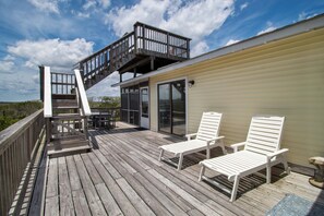 A wooden deck with two white lounge chairs and a table, a glass door leading to a covered area, and a wooden staircase to an upper deck.