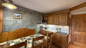 A cozy kitchen with a stone wall, wooden cabinets, a dining table with chairs, and a hanging light fixture. - Quiet 2-bedroom apartment, quick access to slopes, Les Saisies (Les Saisies)
