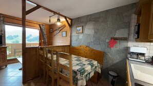 A cozy room with a bed, wooden chairs, a stone wall, a small kitchen area, and a balcony with a view. - Quiet 2-bedroom apartment, quick access to slopes, Les Saisies (Les Saisies)