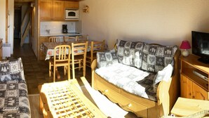 A cozy room with a wooden bed, a small table, and a sofa. There is a kitchen area with wooden cabinets and a microwave.