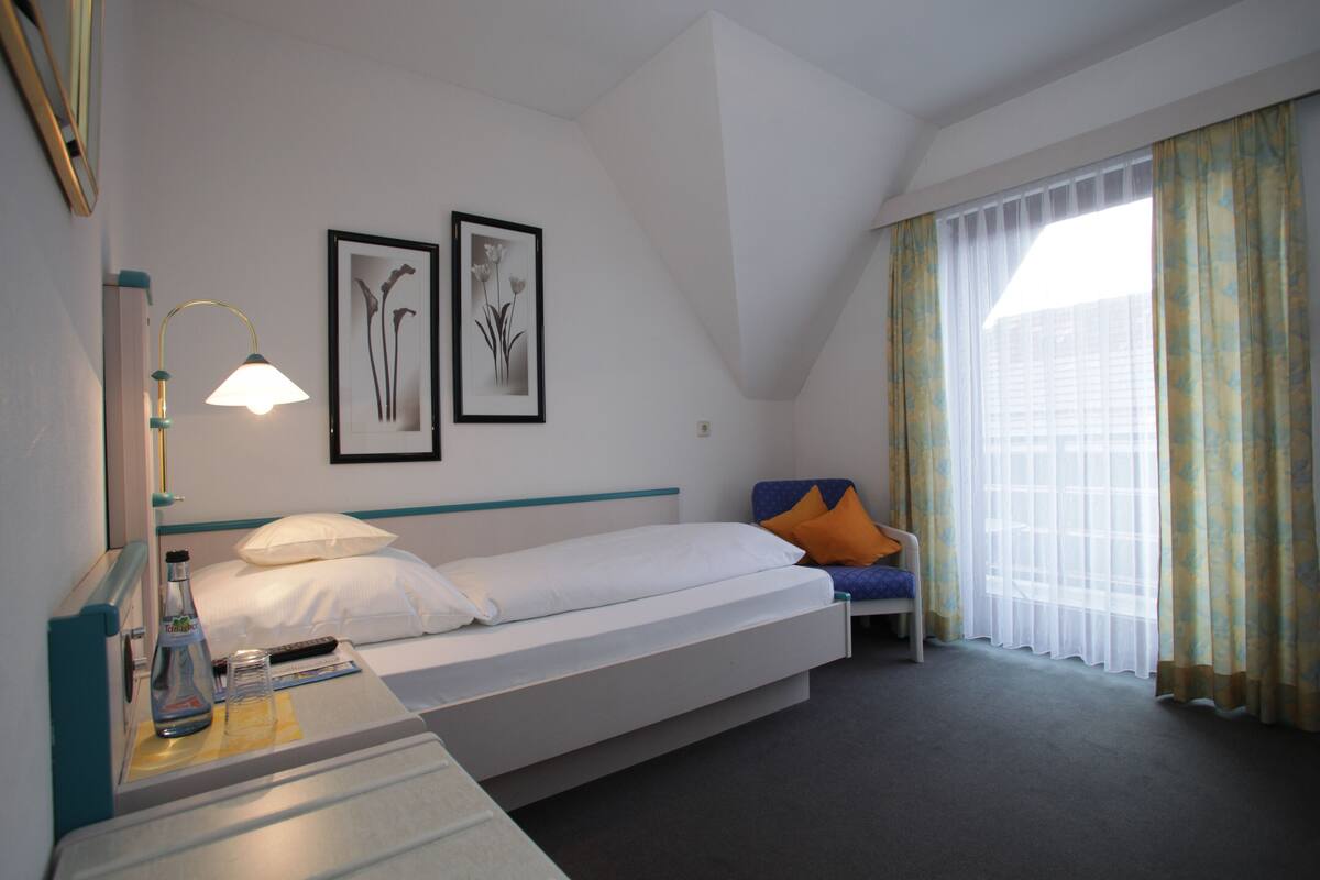 Habitación de hotel con cama, mesita de noche con lámpara, silla y ventana con cortinas.