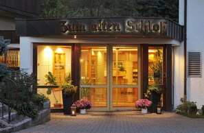 Entrada del hotel Zum Alten Schlof con amplias puertas de cristal, plantas en macetas y un cartel sobre las puertas.