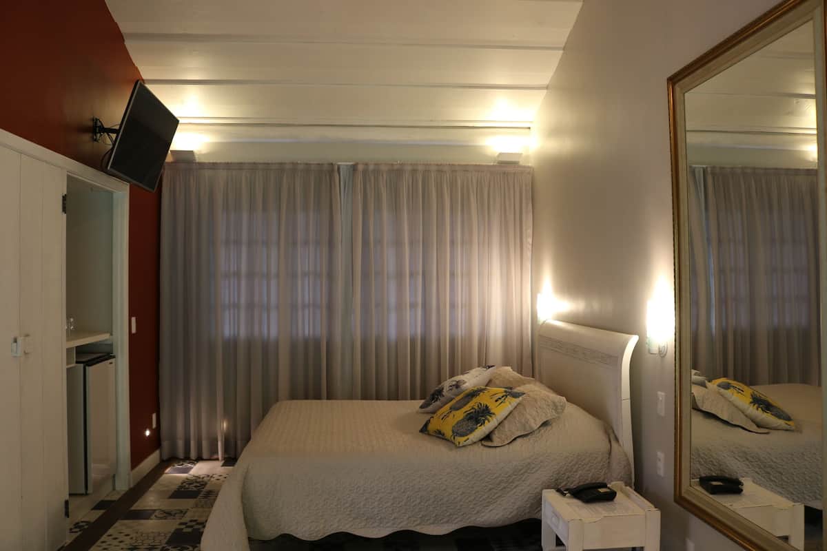 A hotel room with a bed, a bedside table, a mirror, a TV mounted on the wall, and a window with curtains.
