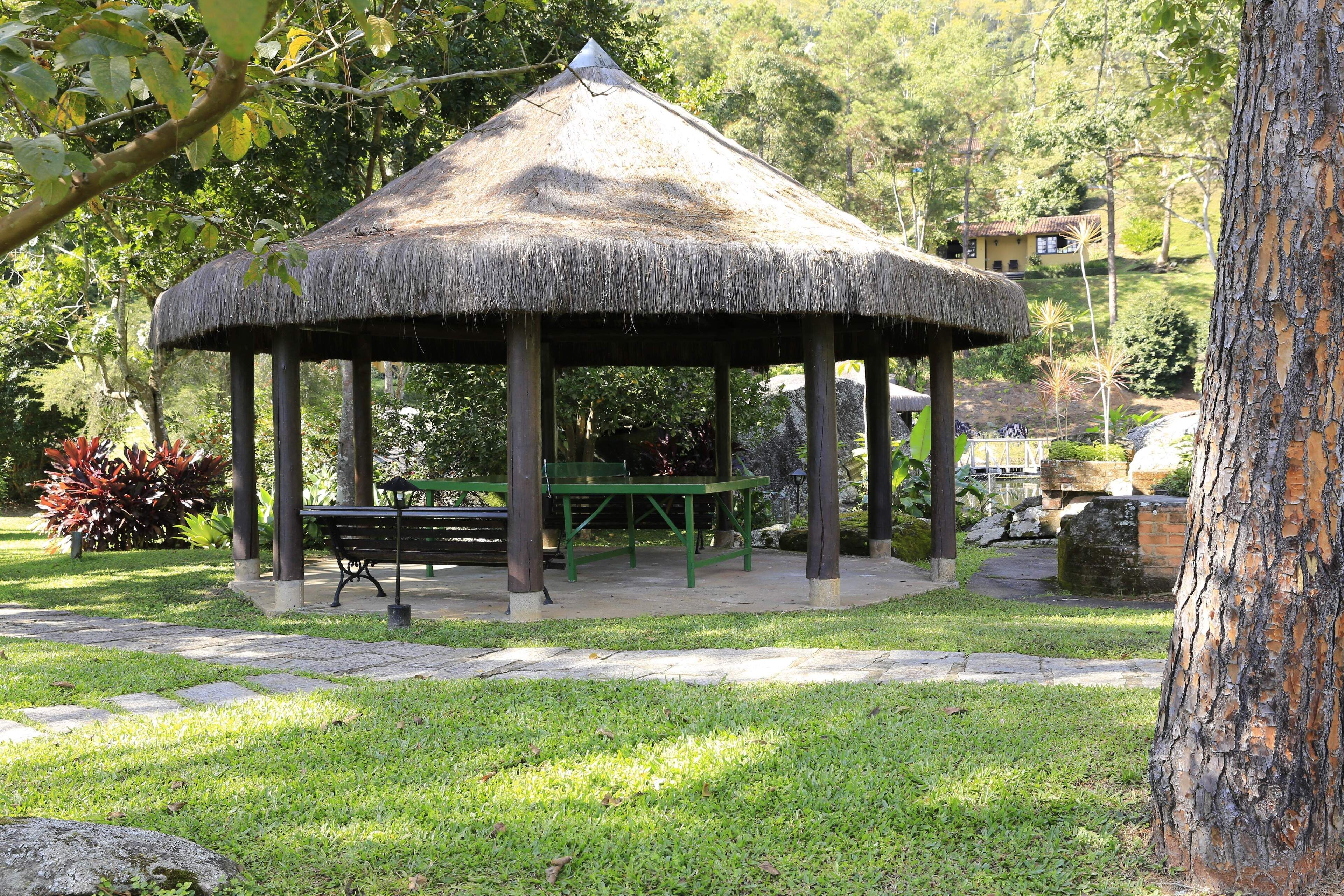 A thatched gazebo with a bench and table, surrounded by grass and trees.