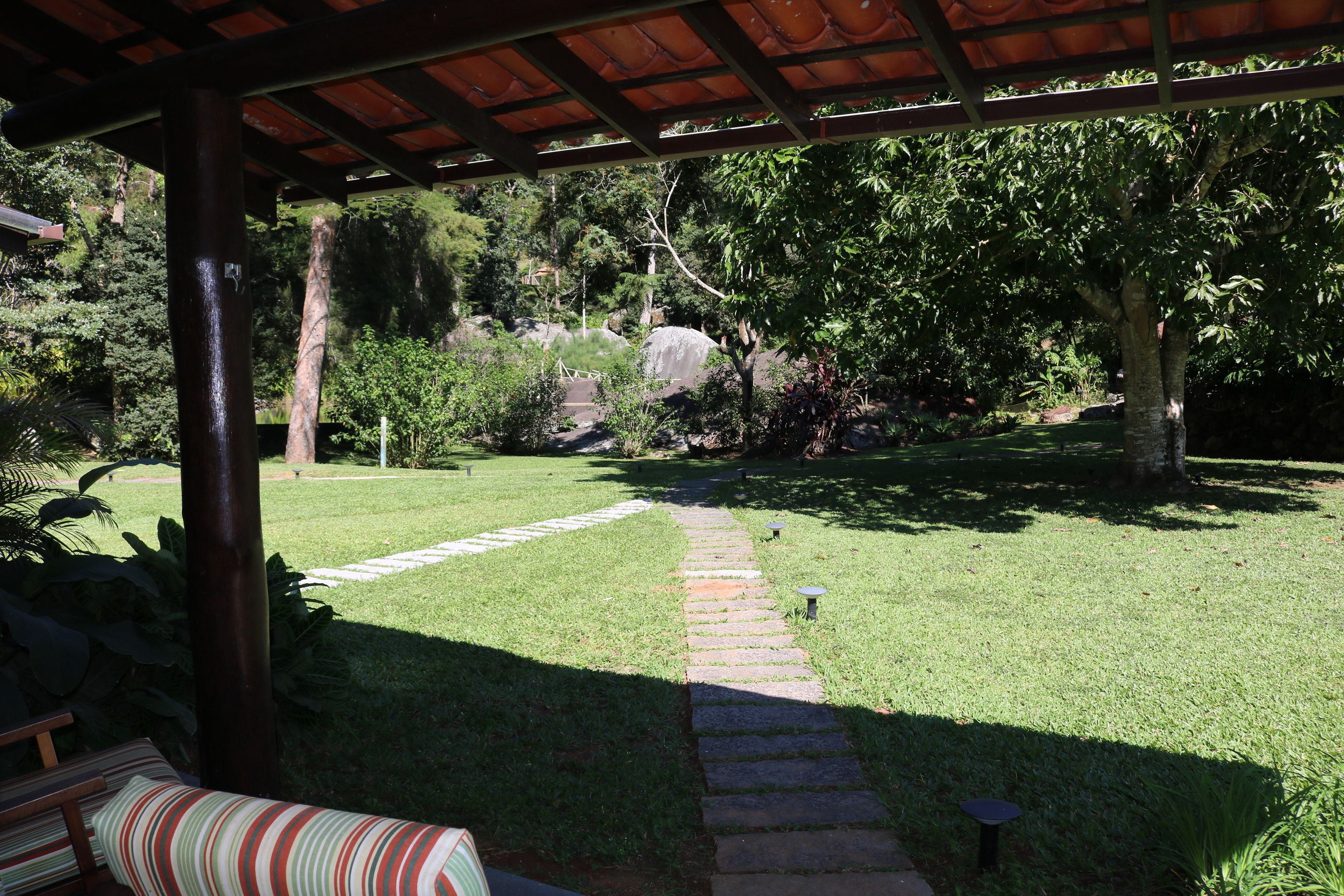 Une terrasse couverte, avec un chemin de pierre menant à une zone herbeuse agrémentée d’arbres et offrant une vue sur une colline.