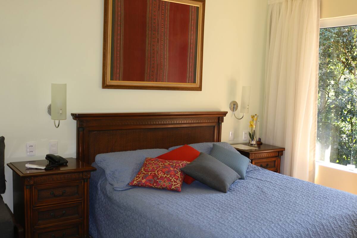 A bedroom with a wooden headboard, a blue bedspread, and two pillows. There is a nightstand with a lamp and a vase of flowers. A painting hangs on the wall above the bed.