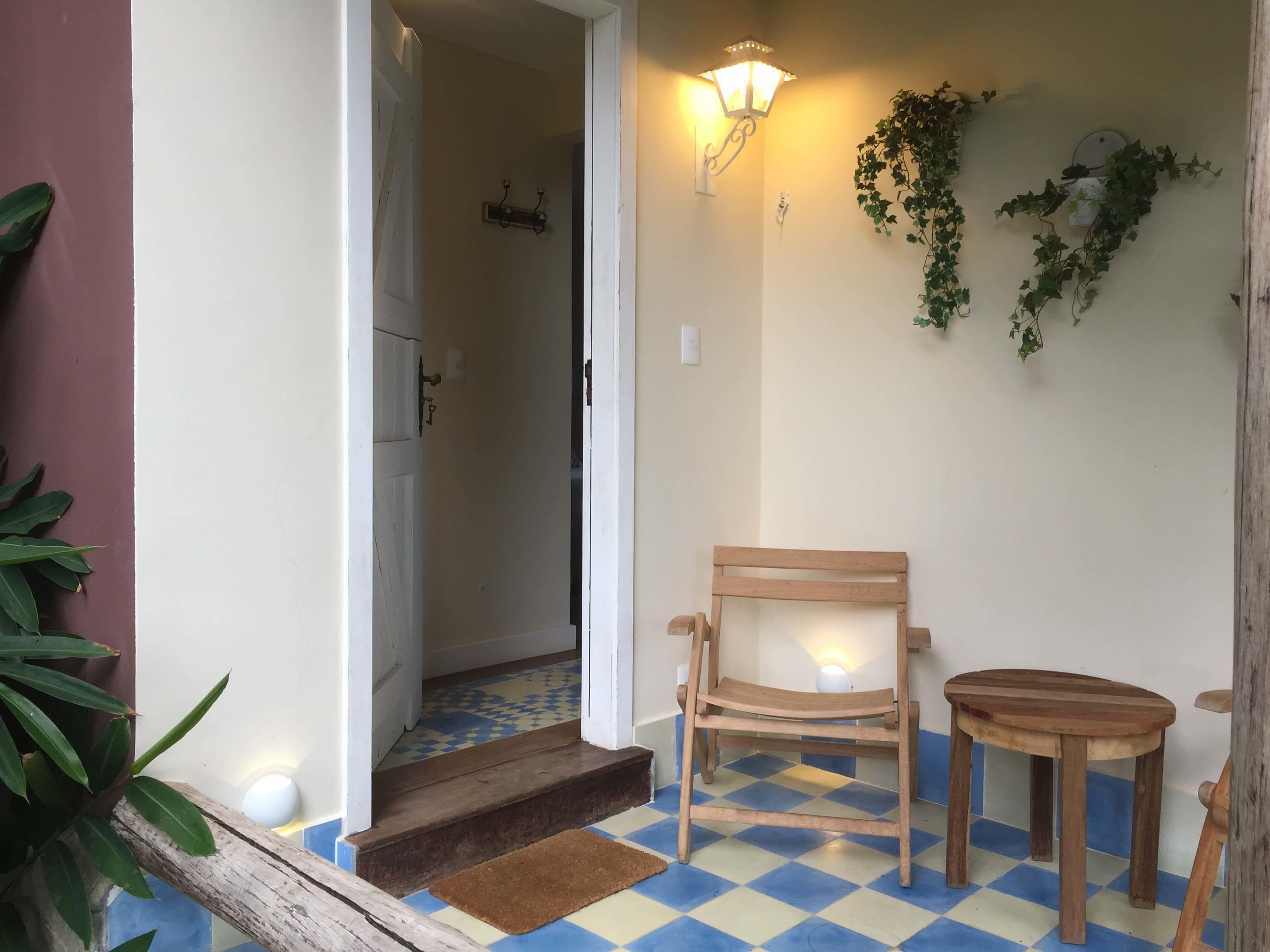 A wooden chair and table outside a building with a checkered floor, a door, and a wall-mounted light fixture.