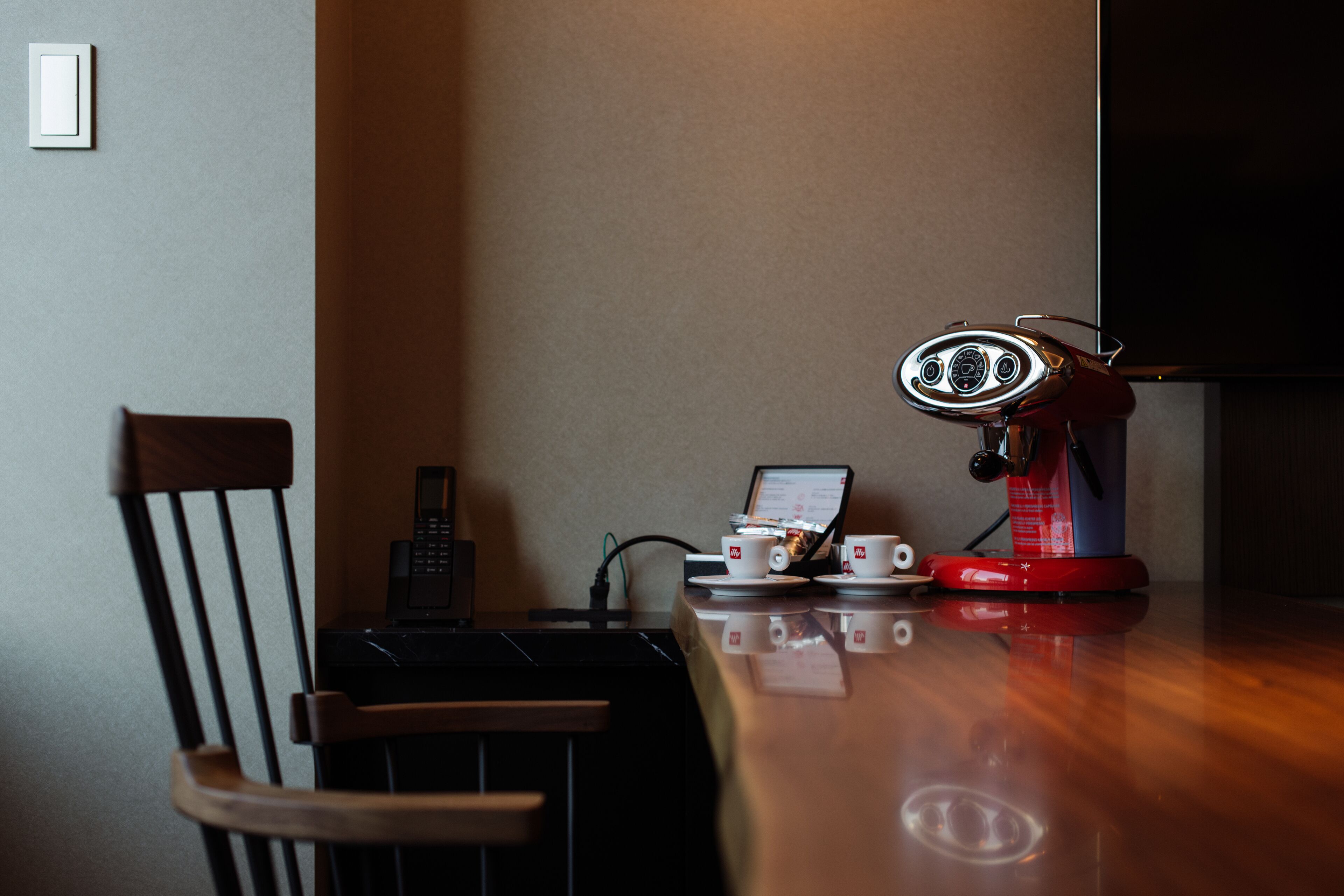 Uma cadeira de madeira, uma mesa com um laptop, uma máquina de café vermelha e um telefone.
