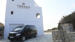A white building with "THEASIS LUXURY SUITES" signage, a black van parked in front, and stone walls. - Theasis Luxury Suites (Naxos)