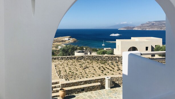 A view through an arched window overlooking a coastal landscape with a clear blue sea, a yacht, and a rocky terrain. - Theasis Luxury Suites (Naxos)