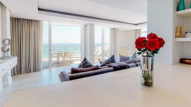 A modern living room with a sofa, a coffee table, and a vase with red flowers. There is a large window with a view of the ocean. - Horizon Bay 1602 (Cape Town)