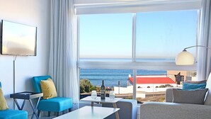 A modern hotel room with a large window offering a view of the sea and buildings. There is a sofa, a small table with glasses and a bottle, and a TV mounted on the wall. - Blouberg Heights 202 (Cape Town)
