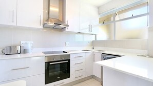 A modern kitchen with white cabinetry, a Bosch oven, a coffee maker, and a window above the sink. - Blouberg Heights 202 (Cape Town)