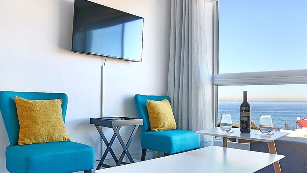 A hotel room with two blue armchairs, a flat-screen TV, a small table with a bottle and glasses, and a view of the sea. - Blouberg Heights 202 (Cape Town)