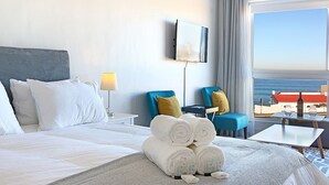 A hotel room with a bed, a TV mounted on the wall, a view of the beach, and a table with a bottle and glasses. - Blouberg Heights 202 (Cape Town)