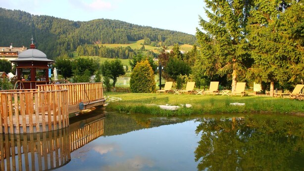 A wooden gazebo with a clock, a pond, and lounge chairs in a park setting. - Alpen Hotel Eghel (Folgaria)