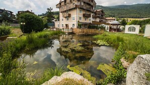 A multi-story building with balconies, a pond with lily pads, and surrounding greenery. - Alpen Hotel Eghel (Folgaria)