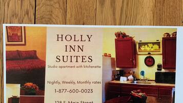 Werbung für Holly Inn Suites: Ein Studioapartment mit Küchenzeile, inklusive eines Essbereichs mit rotem Tisch und Stühlen, einer Küche mit Schränken und einer Mikrowelle sowie einer Wohnbereiches mit rotem Sofa und Kamin.