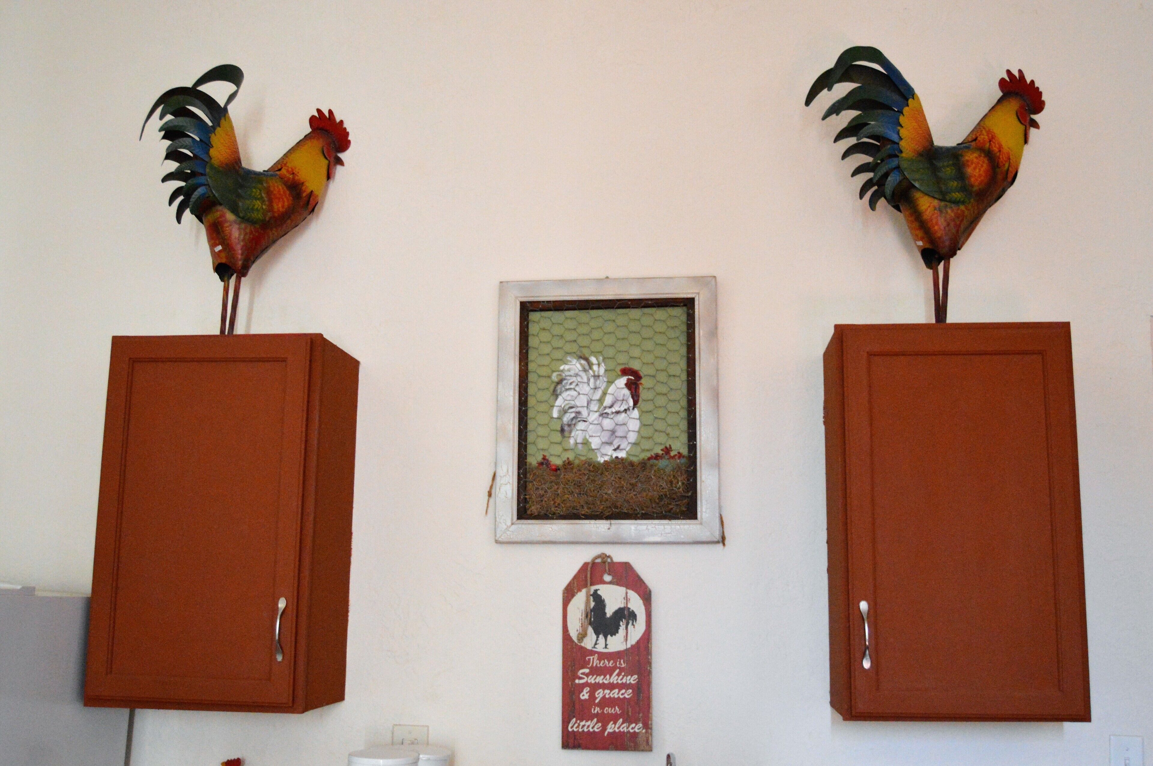 Two wooden cabinets with decorative rooster figures on top, a framed picture of a white rooster, and a sign with a rooster silhouette and text.