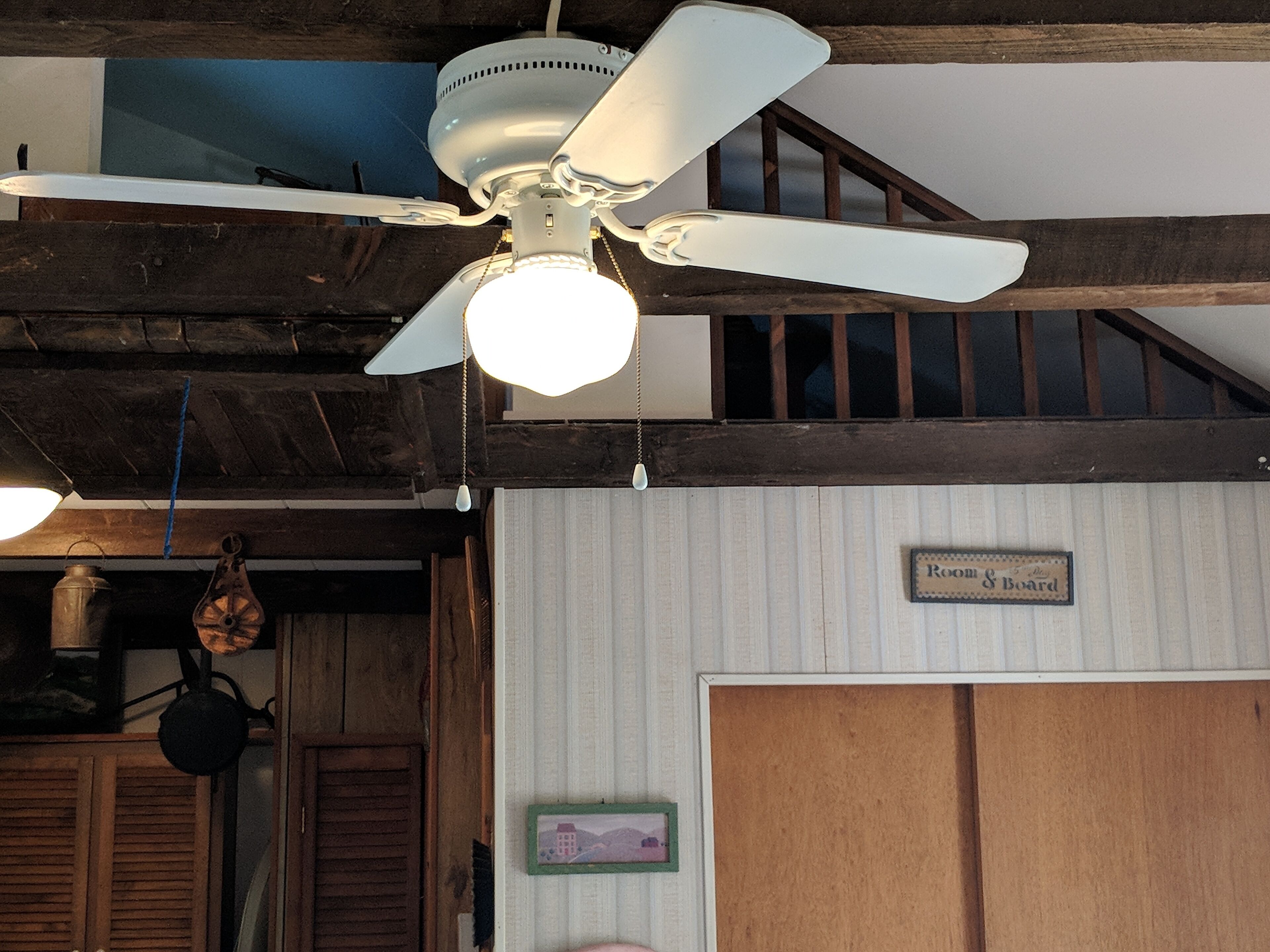 Un ventilador de techo con luces, vigas de madera, una olla de metal y un letrero que dice "Room & Board".