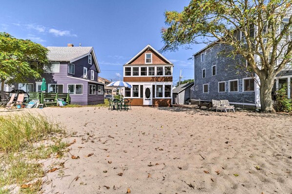 A beachside property with a large house featuring a wrap-around porch, a sandy beach, and a tree.