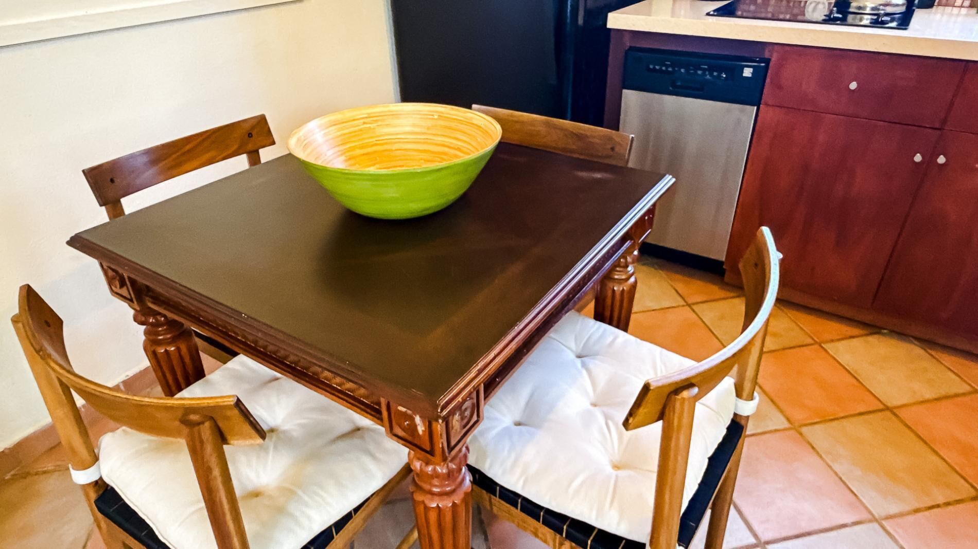 A dining table with a green bowl, wooden chairs with white cushions, and a kitchen area with a dishwasher.