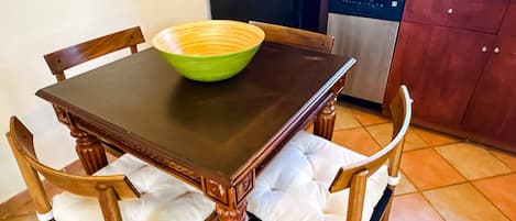 A dining table with a green bowl, wooden chairs with white cushions, and a kitchen area with a dishwasher.