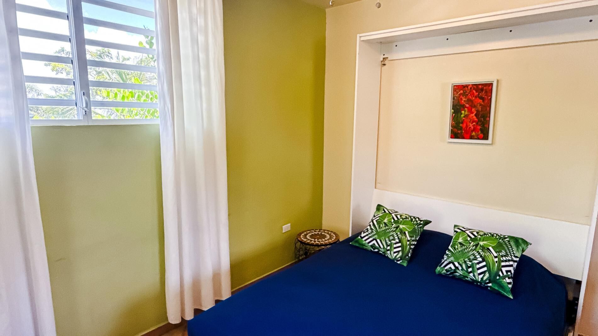 Une chambre à coucher avec un lit, une fenêtre avec des rideaux, une petite table ronde et un tableau encadré accroché au mur.