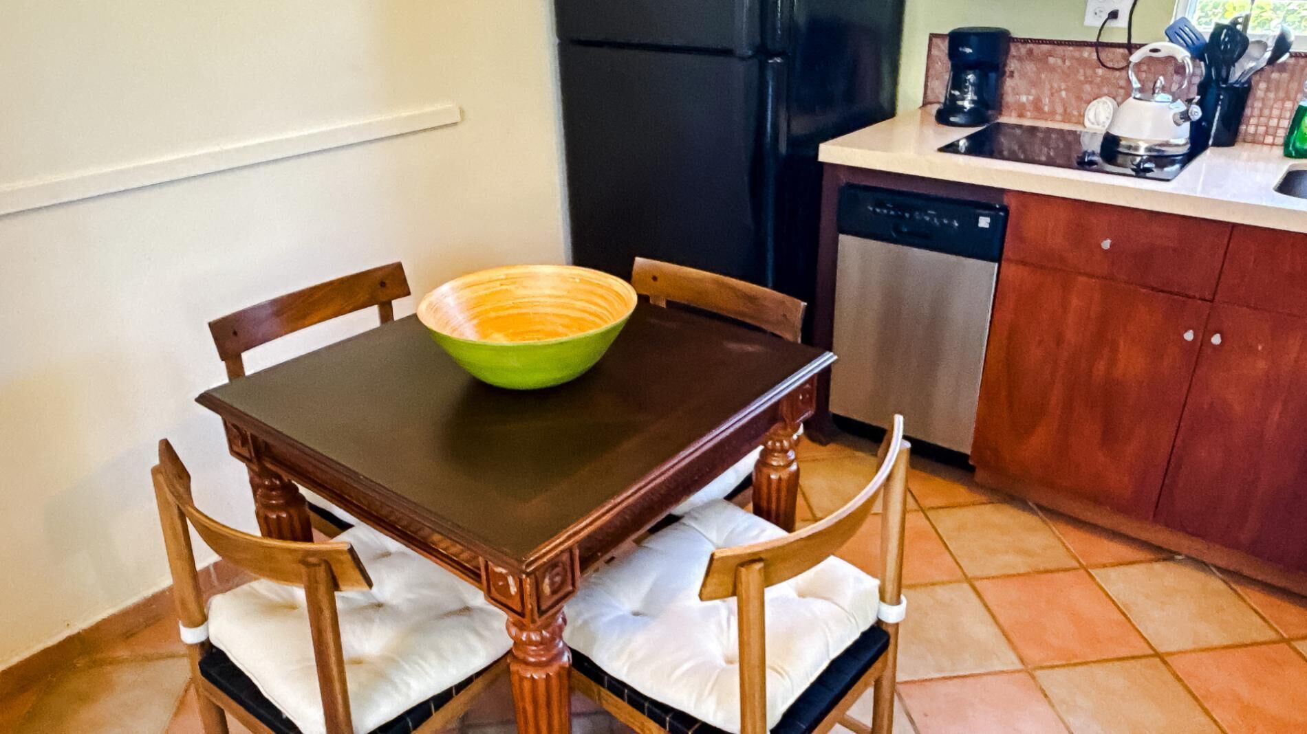 Een eettafel met een groene kom, houten stoelen, een zwarte koelkast en een keukentrap met een koffiezetapparaat.