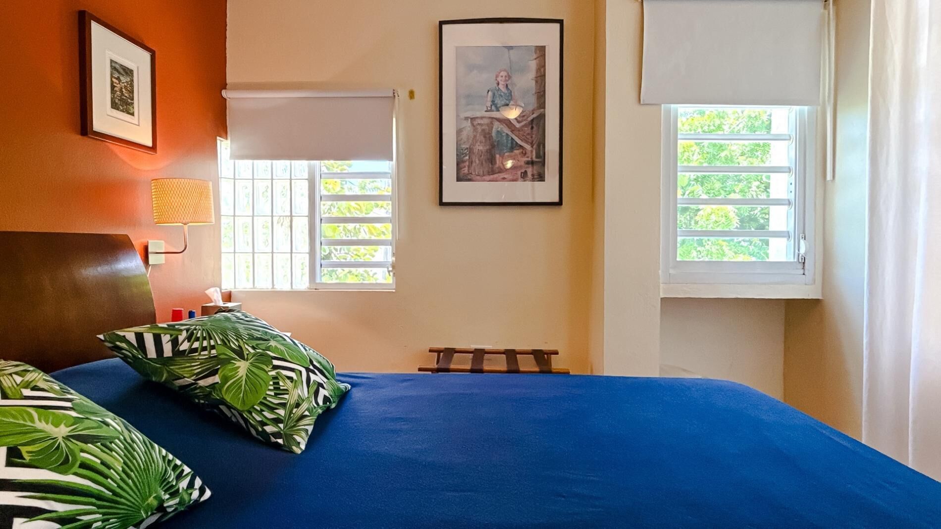 A bedroom with a blue bedspread, two pillows with a tropical leaf pattern, a framed picture on the wall, and two windows with white blinds.