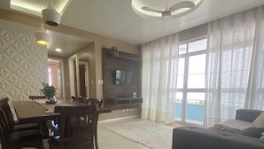 A modern living room with a dining area, a flat-screen TV mounted on a dark wood wall, and a large window with sheer curtains. - Family apartments - air conditioning, fan, internet... fully furnished. (Arraial do Cabo)