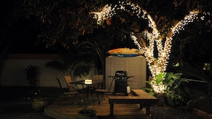 Un patio al aire libre con mesa y sillas de madera, una parrilla y luces colgantes.