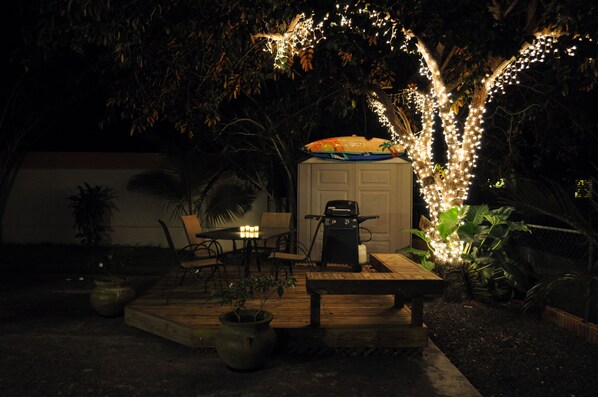 Un patio al aire libre con mesa y sillas de madera, una parrilla y luces colgantes.