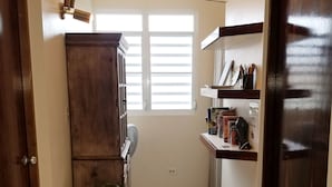 Un pasillo estrecho con un armario de madera, una planta y una estantería con libros y un ventilador.