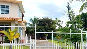 Una entrada cerrada a una propiedad residencial con cerca blanca, camino de entrada pavimentado y abundante follaje, incluyendo palmeras.