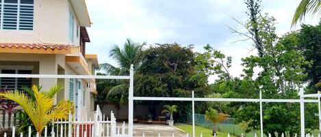 A gated entrance to a residential property with a white fence, a paved driveway, and lush greenery including palm trees.