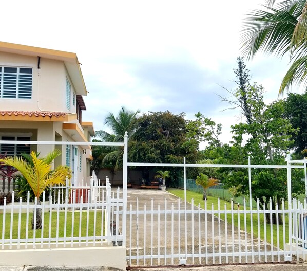 Une entrée fermée par un portail mène à une propriété résidentielle, qui comprend une clôture blanche, une allée pavée et une végétation luxuriante, notamment des palmiers.