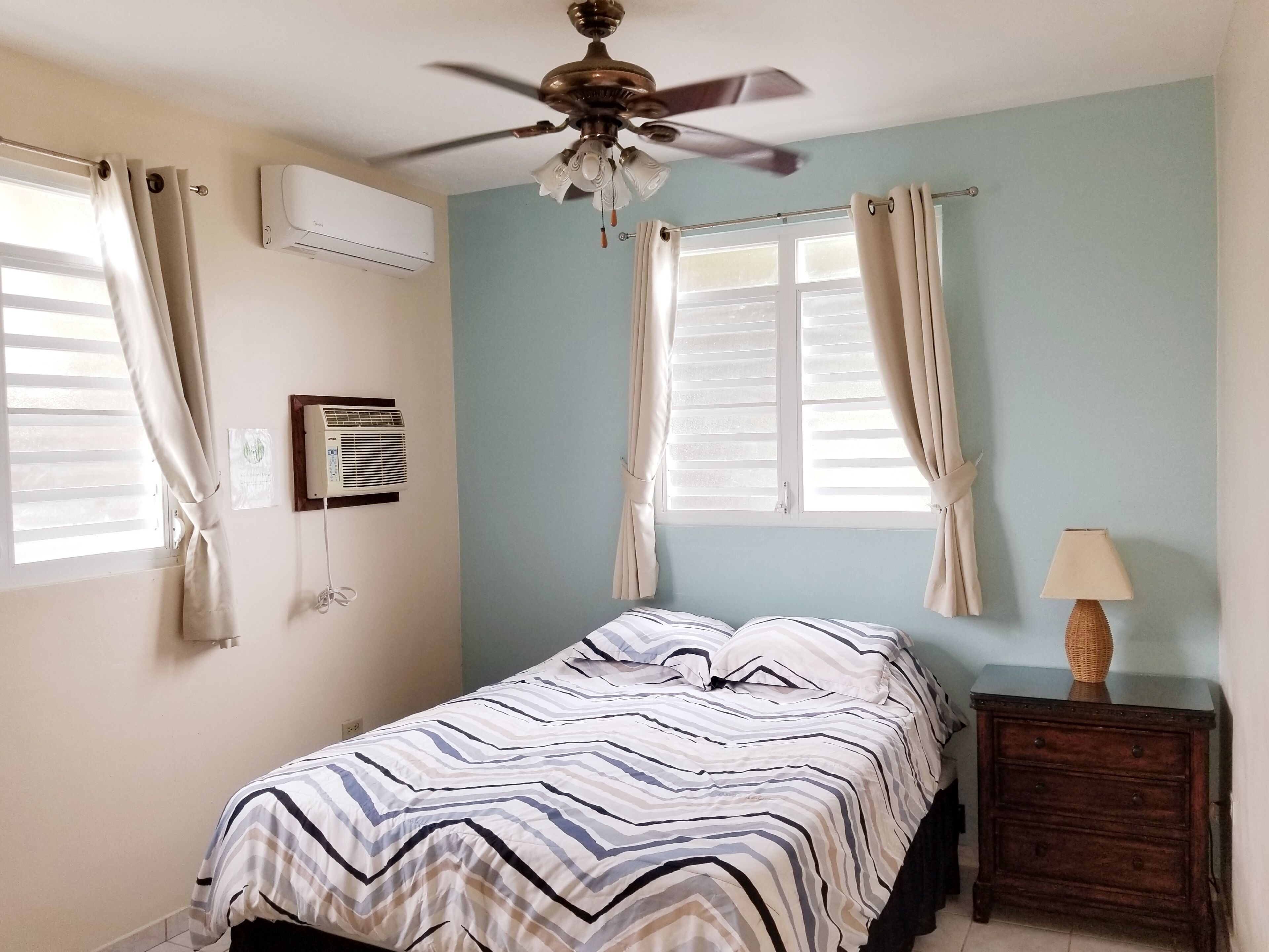 Ein Schlafzimmer mit einem Bett, Deckenventilator, zwei mit Vorhängen versehenen Fenstern, einem Nachttisch mit Lampe und einer Klimaanlage.