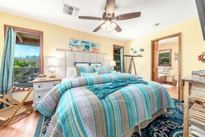 Ein Schlafzimmer mit Bett, Deckenventilator, Nachttisch, Kommode und Fenster mit Vorhängen.