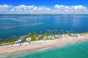 Vista aérea de una zona costera con playa arenosa, un muelle que se adentra en el agua y varios edificios de aspecto similar a un complejo turístico.